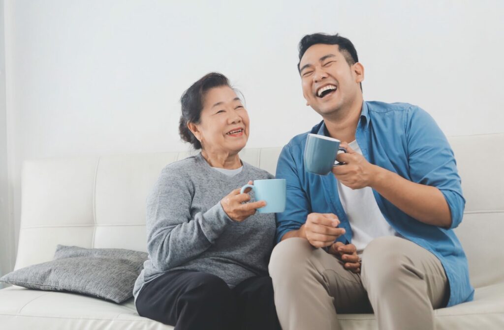 An older adult woman and a her son sitting sitting on a couch, smiling and talking to each other while holding a warm beverage