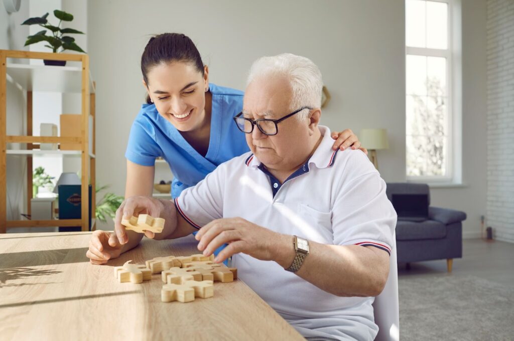 A staff member helps an older adult complete a wooden puzzle on a table in memory care.