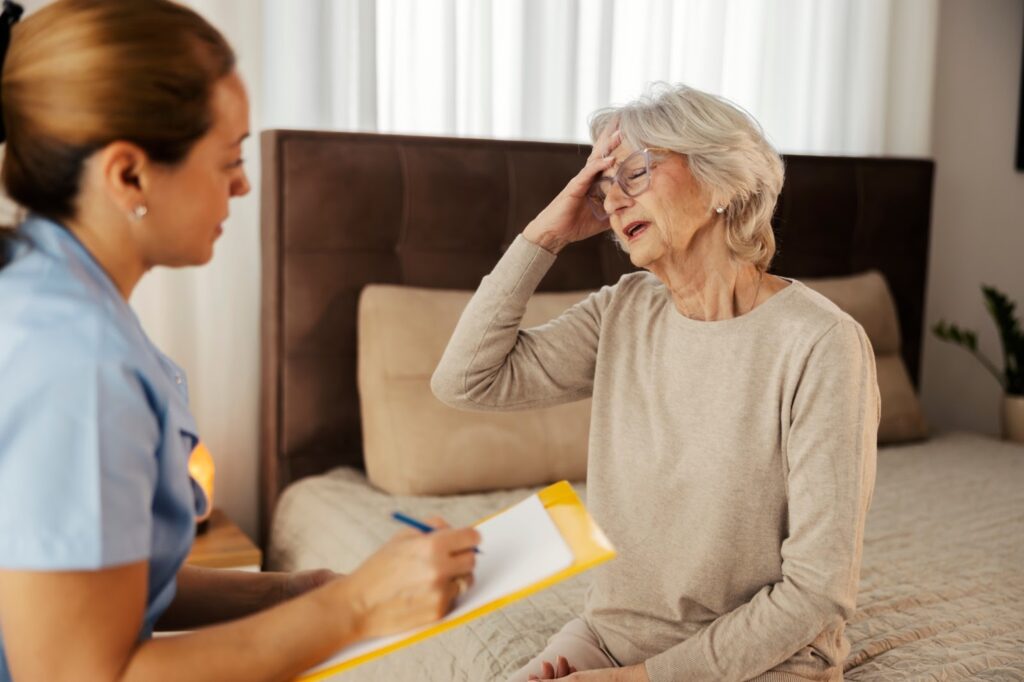 A senior woman clutching her forehead in pain while discussing symptoms to a memory care team member.