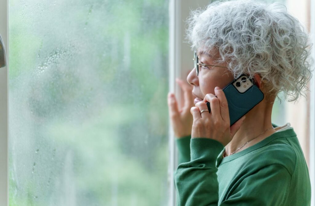 Older adult looking out rain-covered window while talking on the phone during storm