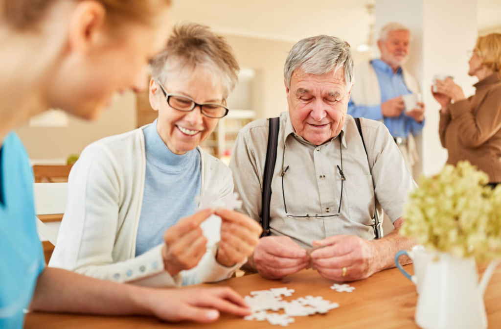 Two older adults assemble a puzzle in the common area of a memory care community with their caregiver's help while two other residents drink tea in the background