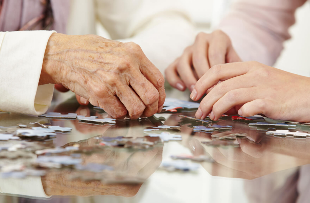 Two older adults assemble a puzzle in the common area of a memory care community with their caregiver's help while two other residents drink tea in the background