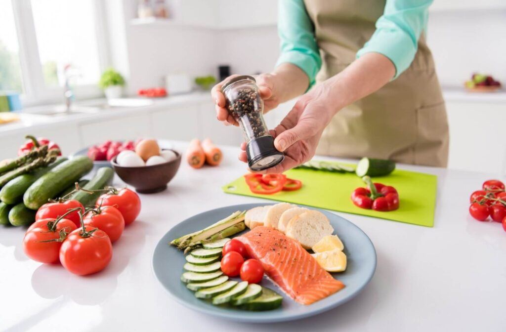 An adult adds pepper to a plate of salmon, cucumber, asparagus, and lemon.