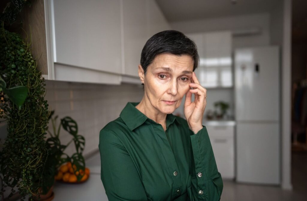 An older adult stands in a dim kitchen and rubs their face in frustration due to their memory loss