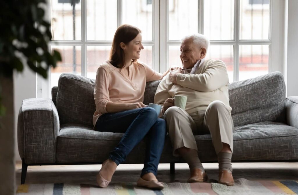 An adult child rests their hand on their older parent's shoulder and smiles during a warm conversation about memory loss in a living room in memory care