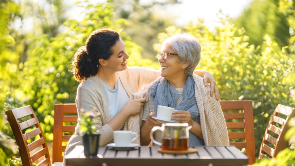 An older adult and their adult child sit at a patio table outside in a garden and sip tea together on a sunlit day