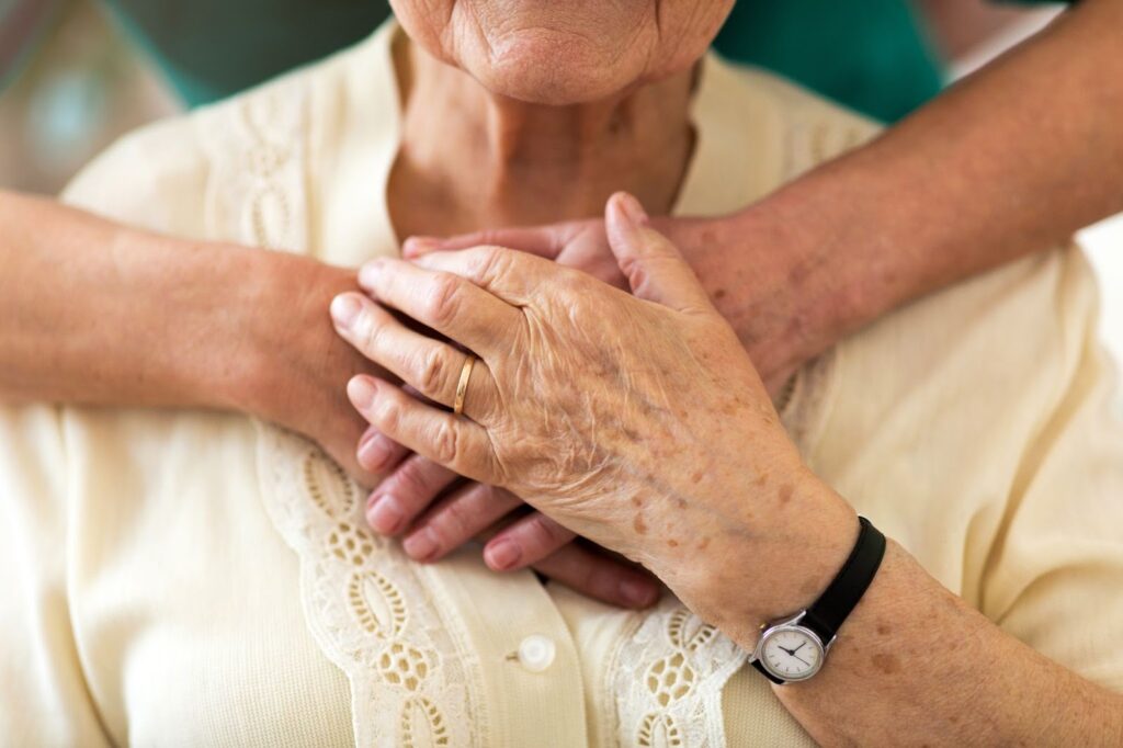 A caregiver gently holds a senior from behind during a quiet, respectful moment together